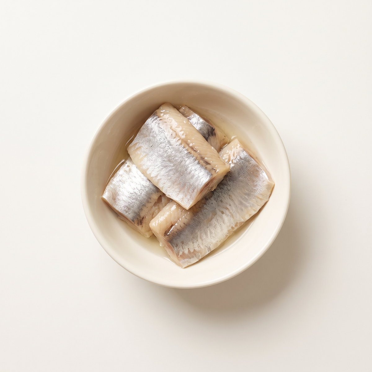 four marinated herring fillet pieces, pearly silver skin and pale beige flesh, gentle sheen of clear marinade, in a small plain ivory bowl