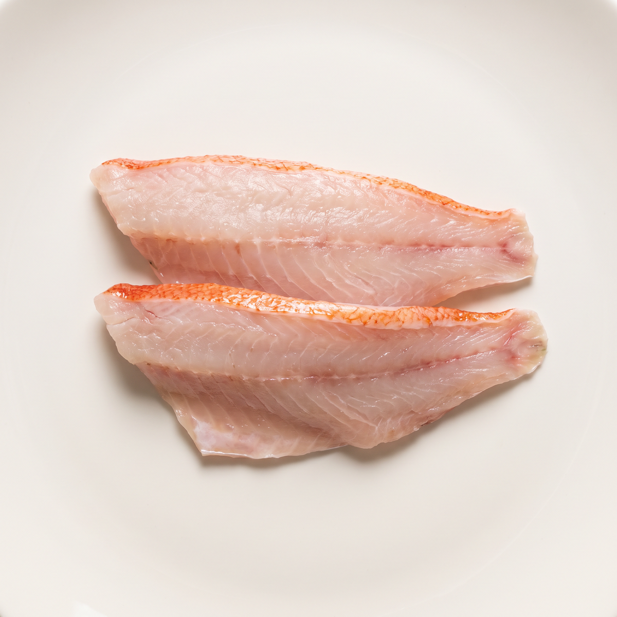 two raw ocean perch fillets, pearly white pale pink flesh, thin vivid reddish skin edges, slender tapered shapes