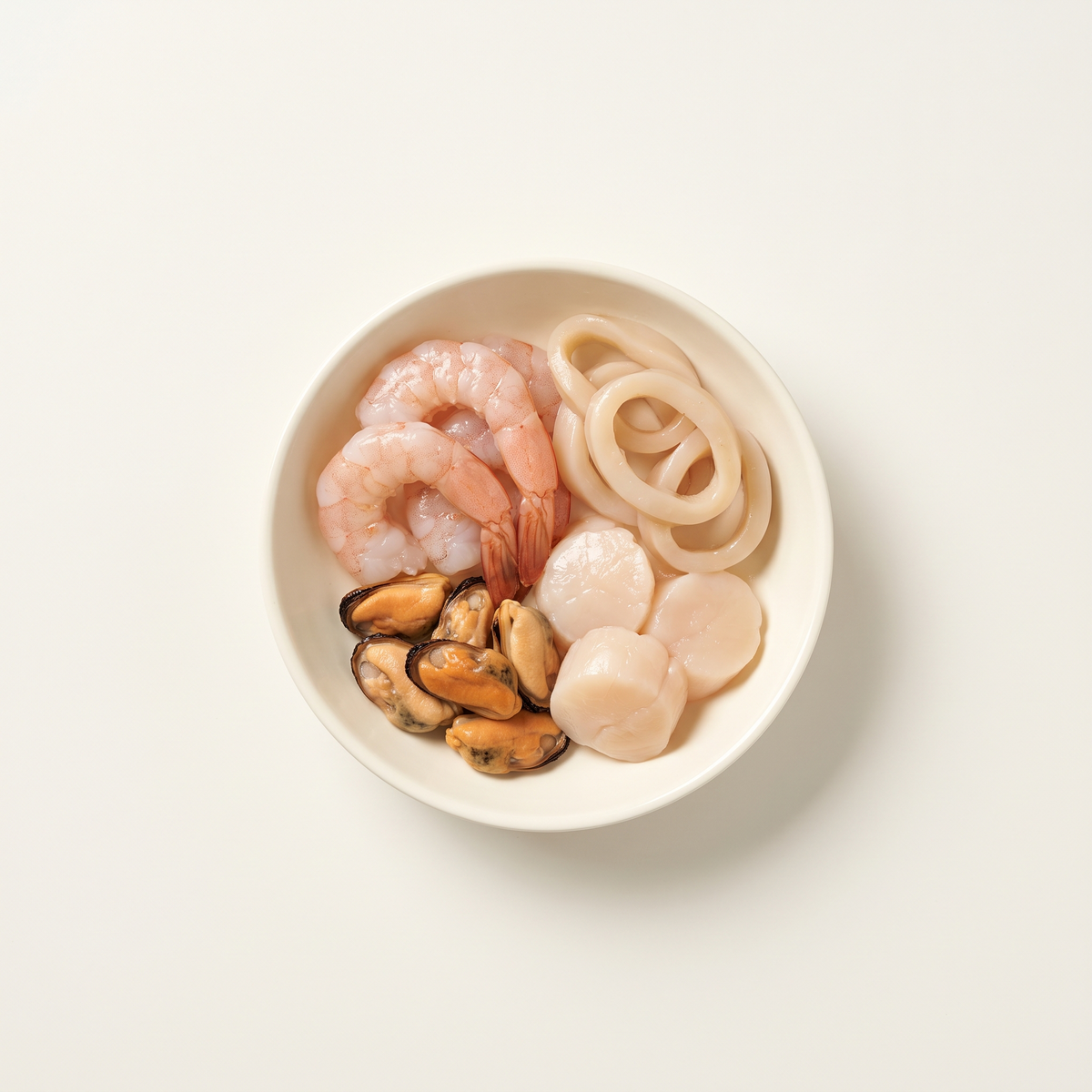 a compact small assortment of peeled raw shrimp, ivory squid rings, mussel meat and raw scallop pieces in a shallow ivory bowl, all four components clearly distinct