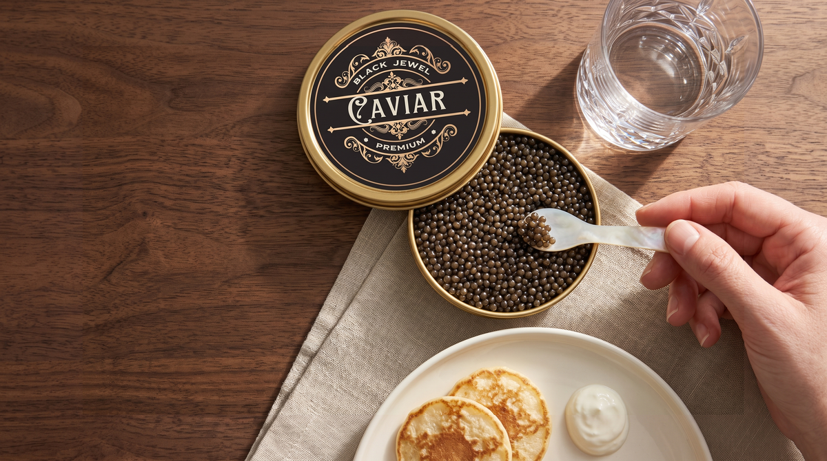 Complete Black Jewel caviar tin and matching ornate lid viewed from above, with the original spoon, hand, crystal glass and blini on walnut